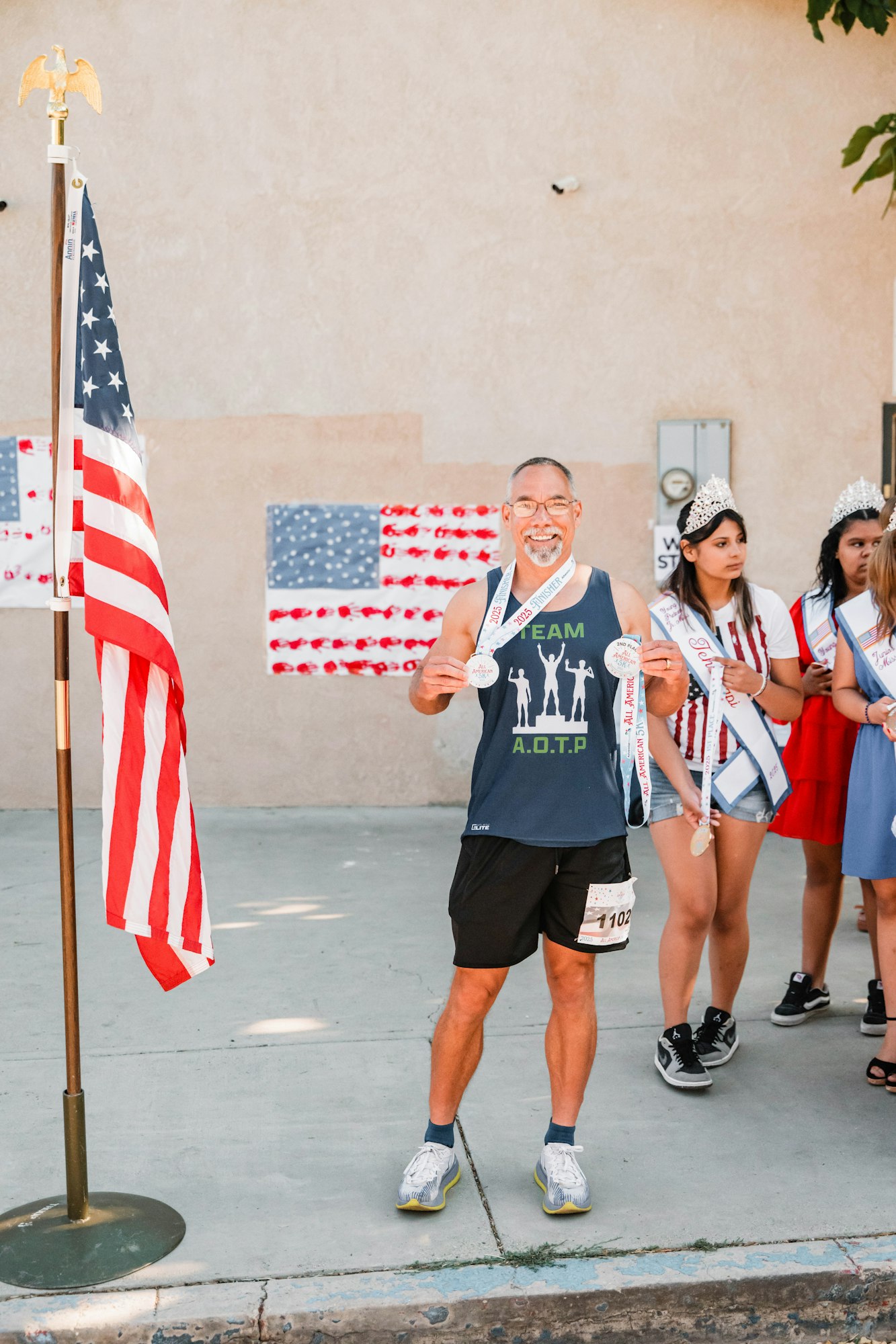 A man smiling with medals, standing by an American flag; people with sashes and tiaras in the background.