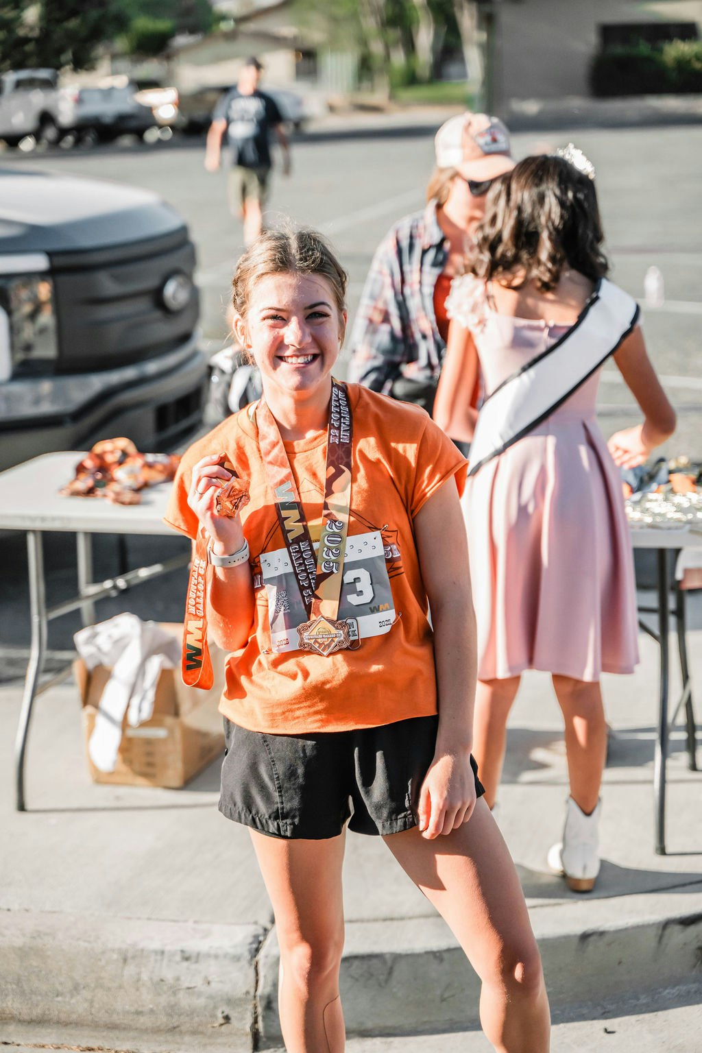 A person in an orange shirt, wearing medals, smiling, with others and a table in the background.