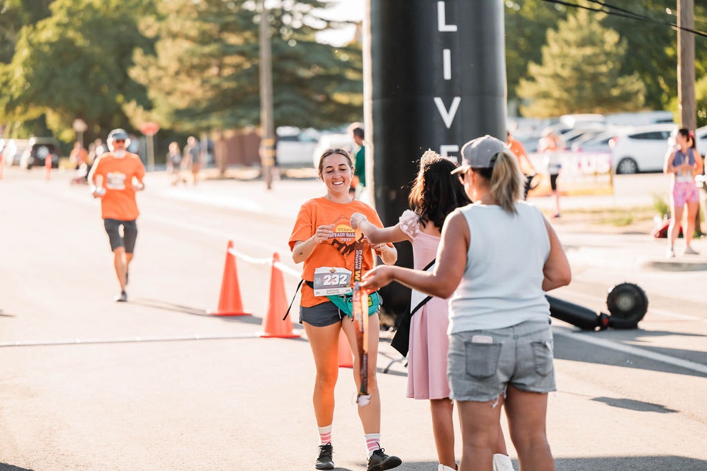 Runner finishing a race, smiling, with people congratulating them. Orange shirt, number 232, outdoor event.