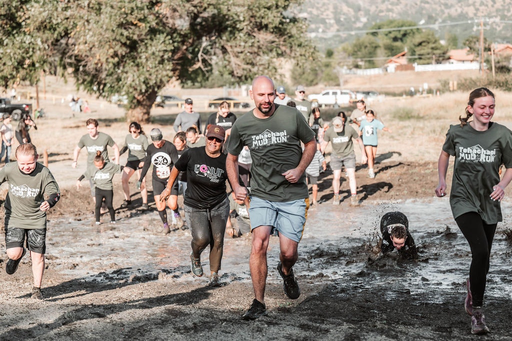 A group of people participating in a muddy outdoor race, smiling and running through a muddy area.