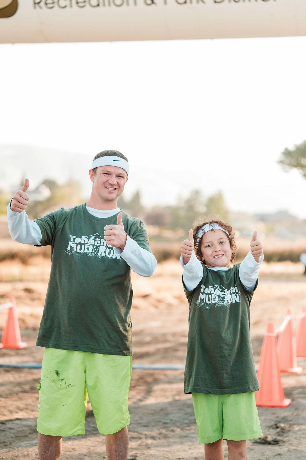 Two people in matching "Tehachapi Mud Run" shirts and headbands giving thumbs up, with orange cones in the background.