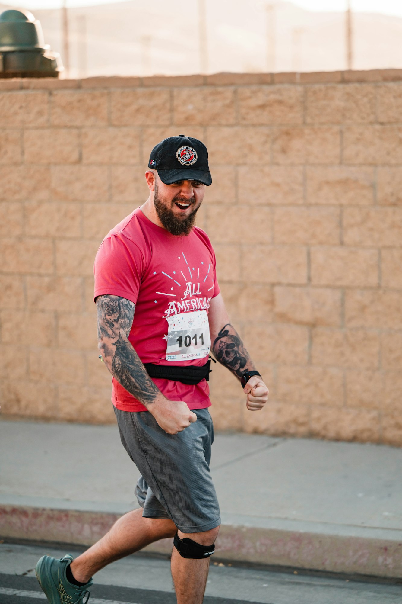 A man with tattoos in a red shirt, wearing a cap, walking confidently with a race bib numbered 1011.