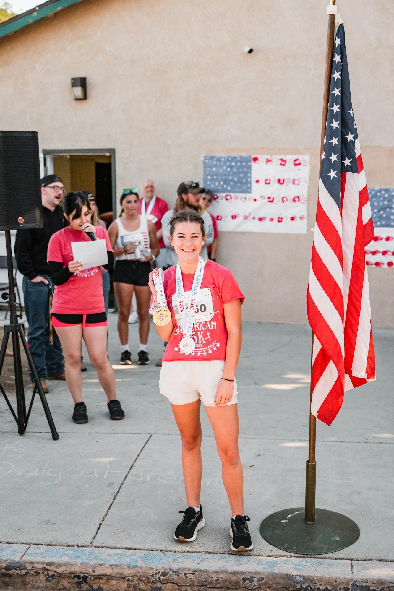 A person smiling, holding medals at an event near a U.S. flag with spectators and a speaker in the background.