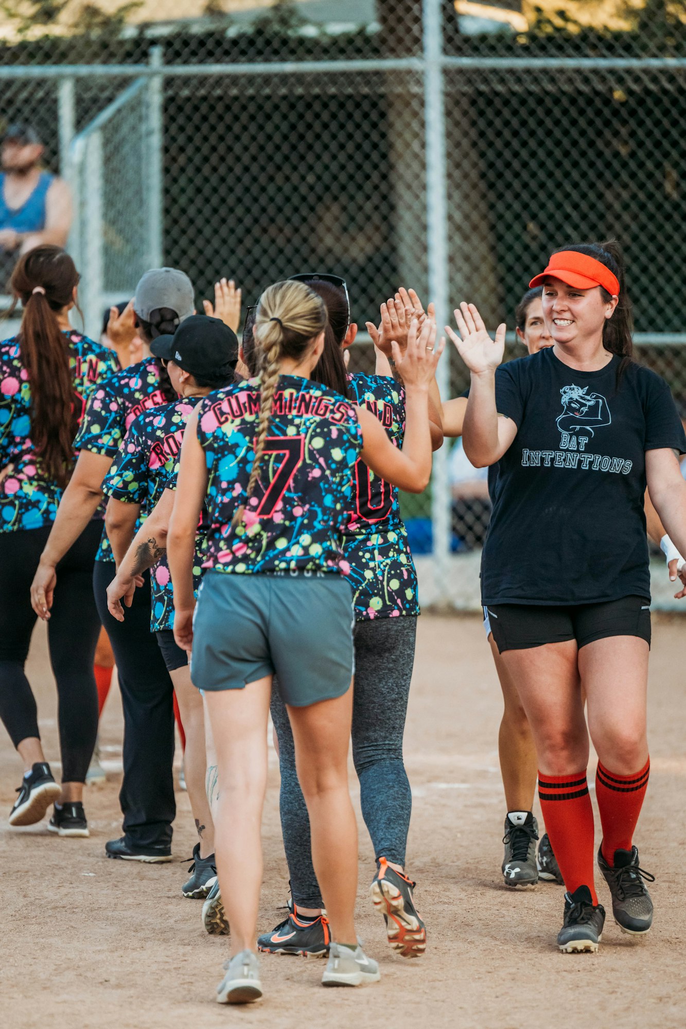 Softball players high-fiving after a game, with one smiling at the camera. They wear colorful jerseys and sports attire.