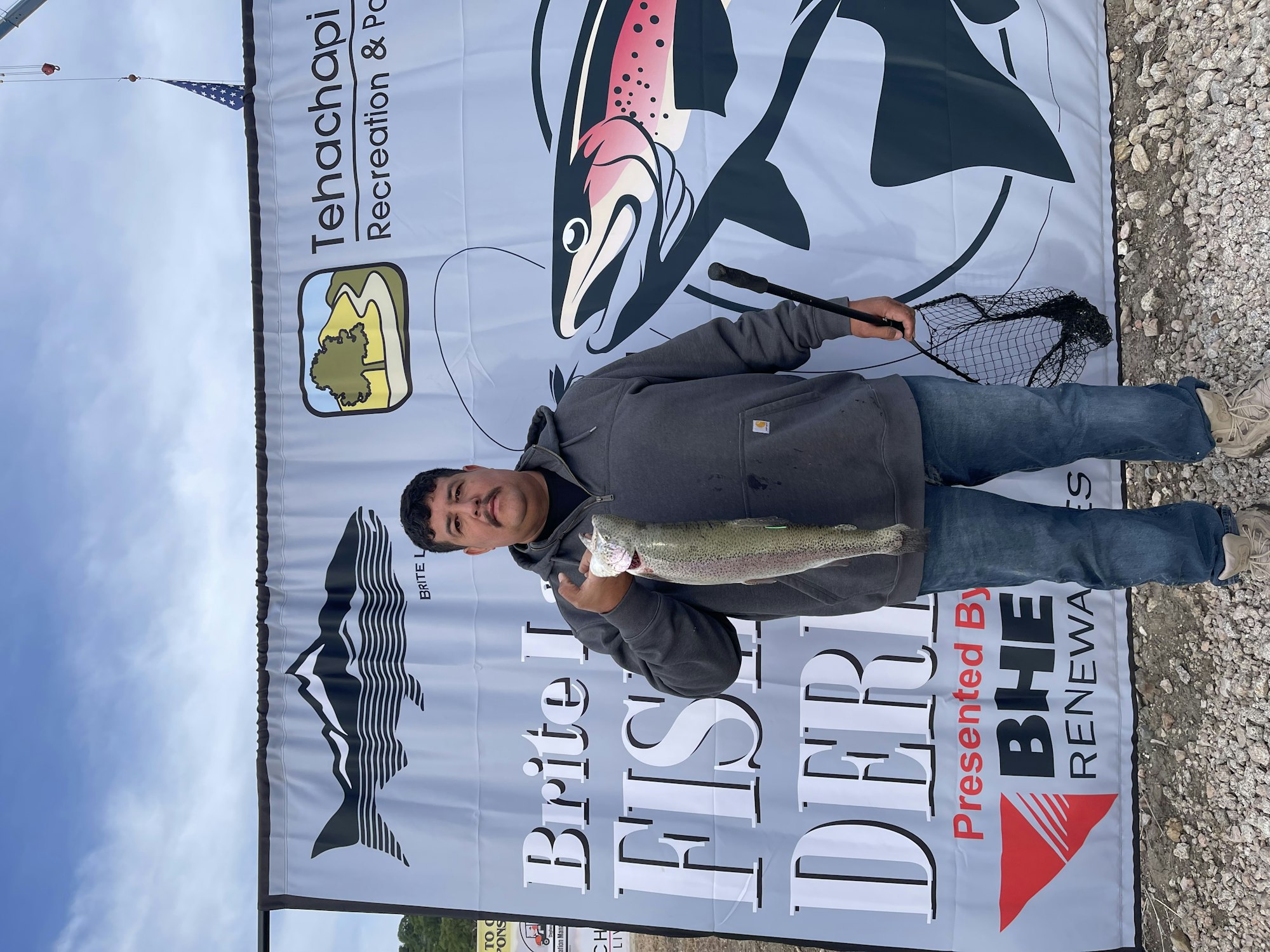 A person holding a fish stands in front of a sign for the Brite Lake Fishing Derby.