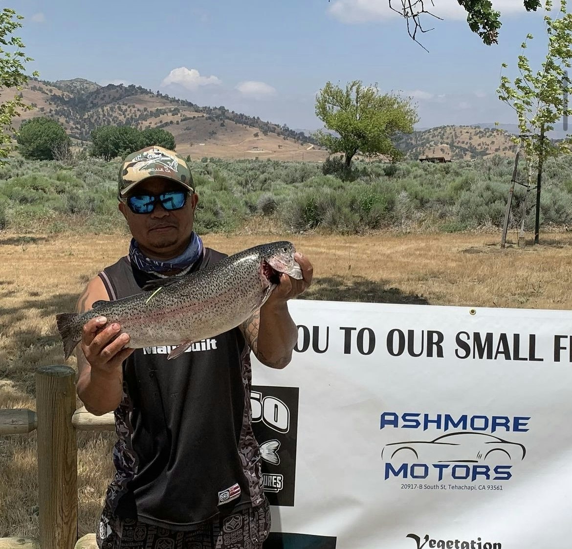 A man in sunglasses holding a large fish with hills in the background and a sponsor banner in the foreground.