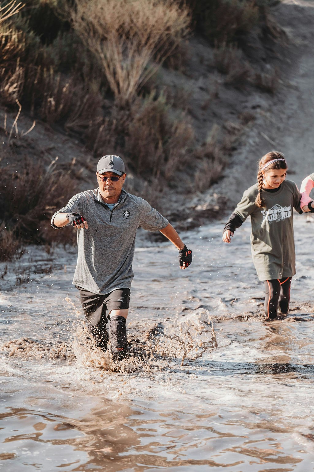 Two people, one adult and one child, are running through muddy water in an outdoor setting.