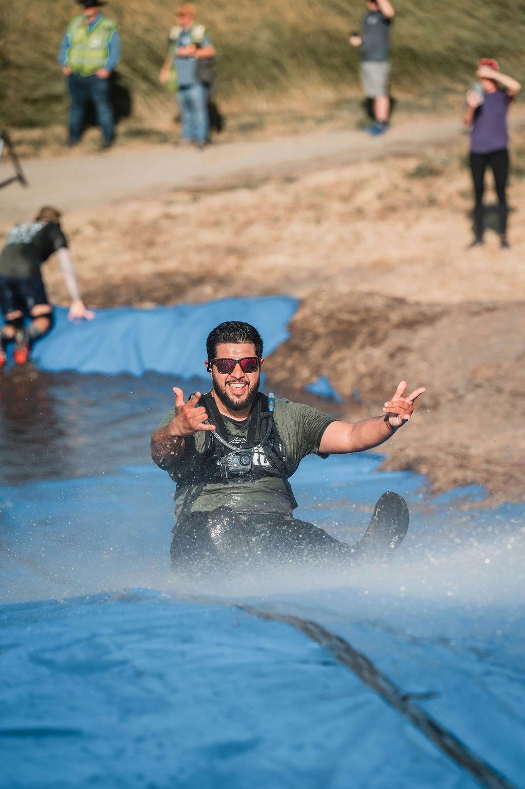 A man sliding on a wet inflatable, smiling, making hand gestures, with several people watching in the background.