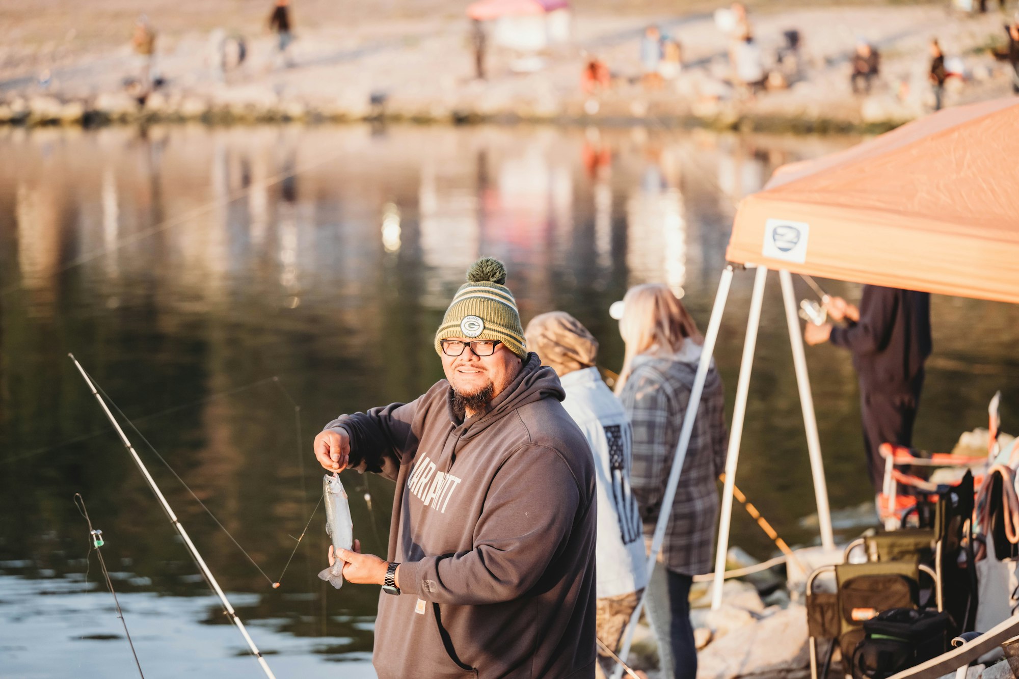 A person smiling while holding a fish at a lakeside, with fishing gear and people in the background.