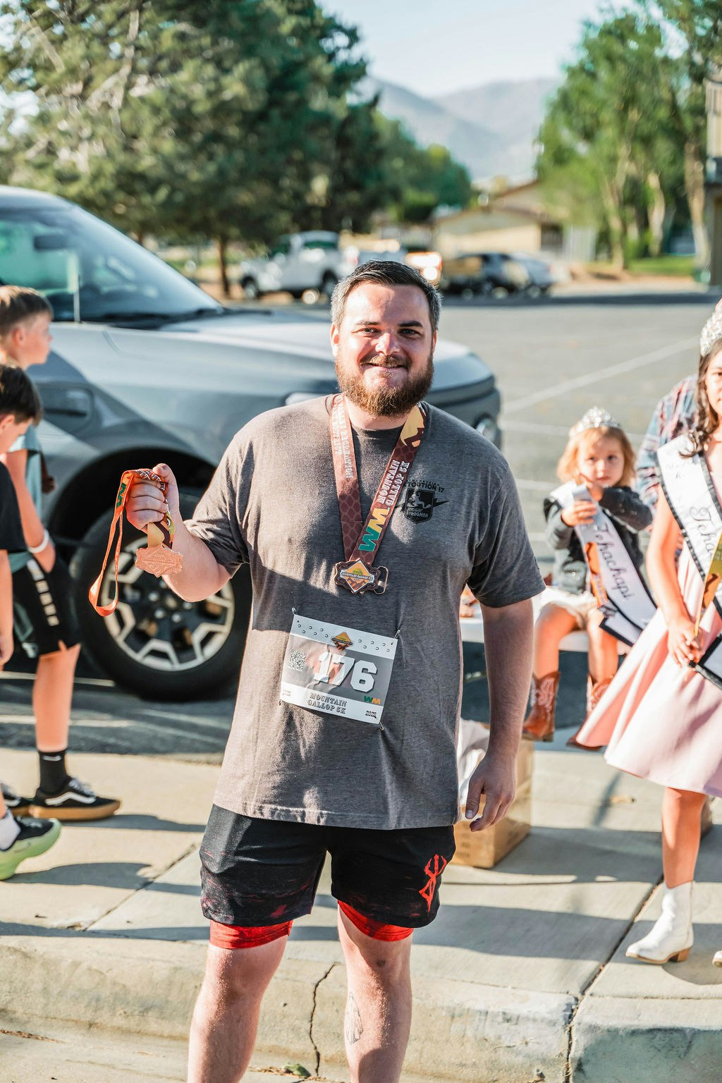 A man with a race number and medals standing outdoors, smiling. Children are in the background, some wearing sashes and crowns.