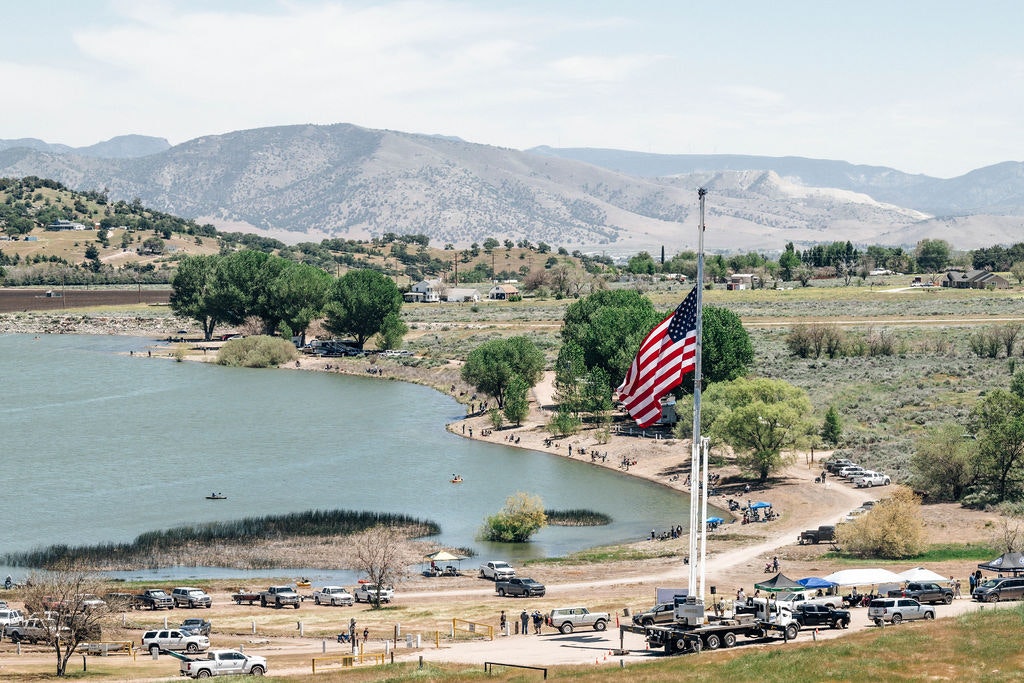 A lakeside scene with an American flag in the foreground, vehicles parked, people enjoying water activities, and mountains in the background.