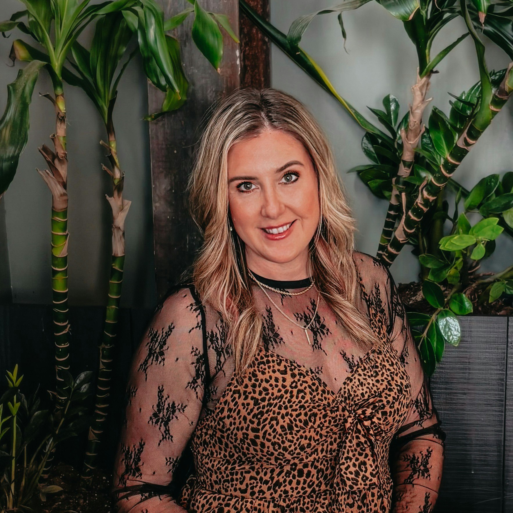 A smiling woman with long hair poses against a backdrop of indoor plants, wearing a leopard print dress with lace details.