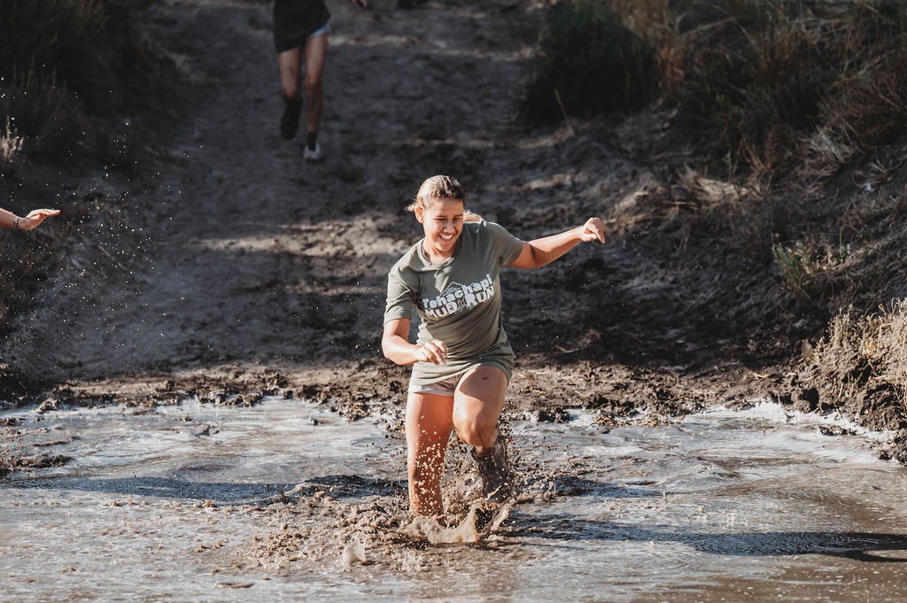 Person running through a muddy obstacle course, splashing water, with a determined expression.