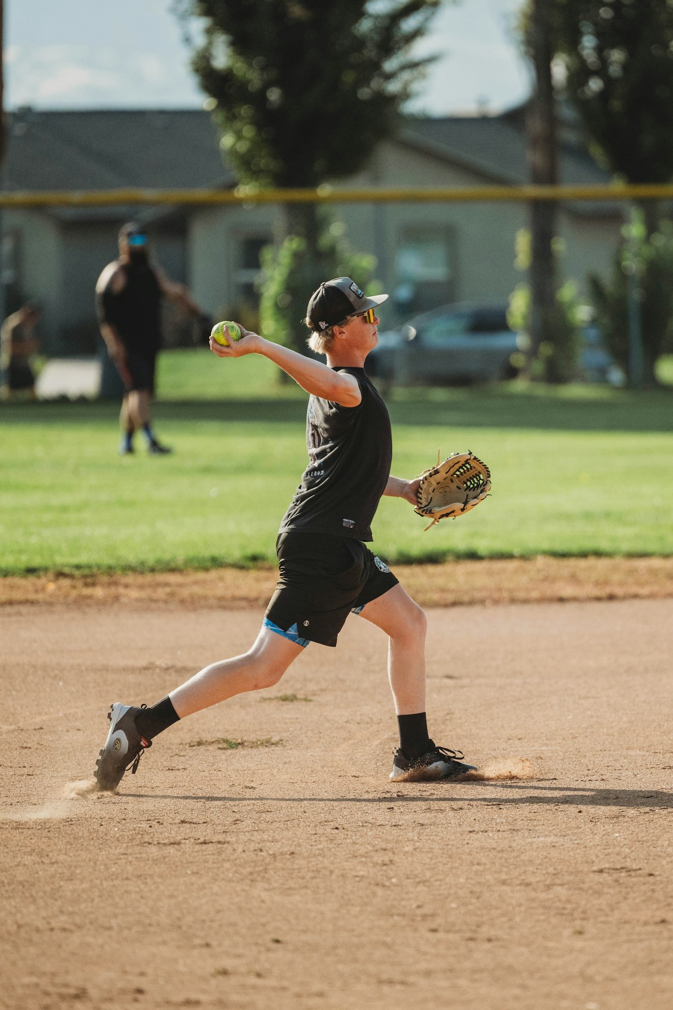 A young player throwing a softball on a sunny field, with another person in the background.