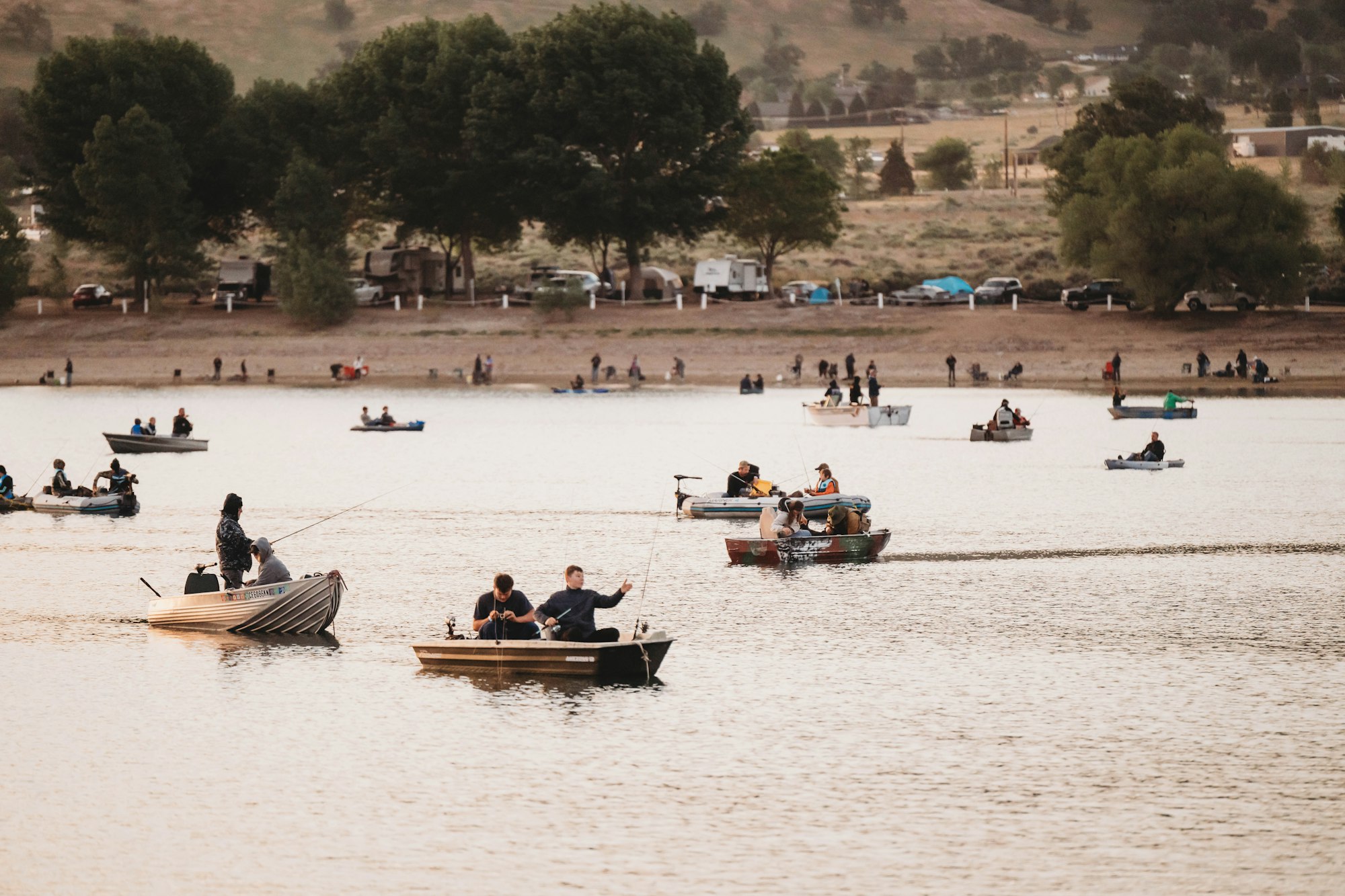People fishing on boats in a calm lake, with a tree-lined shore and several parked vehicles and tents in the background.