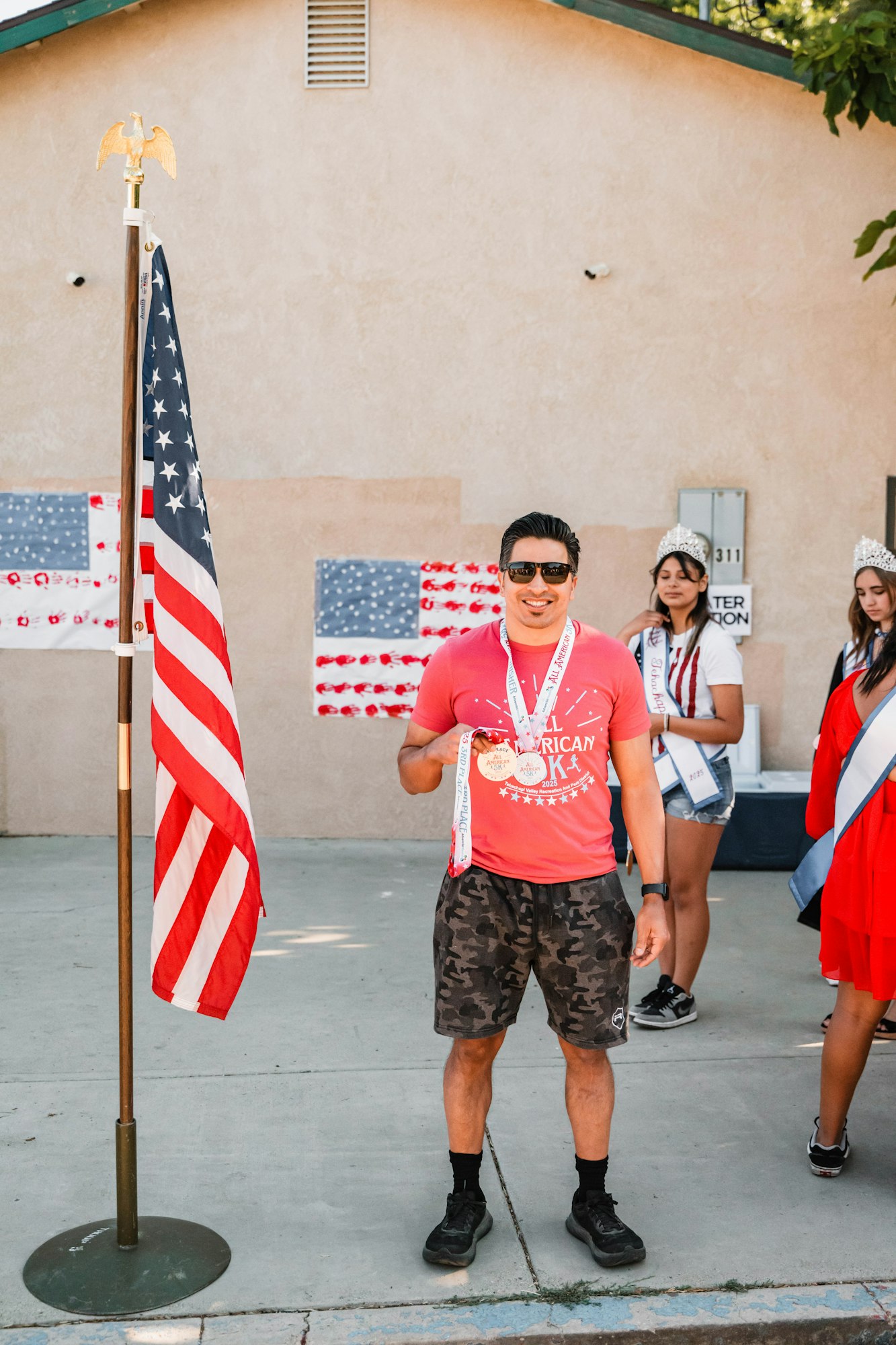 Man with medals in front of a U.S. flag, wearing sunglasses, near people with sashes and crowns.