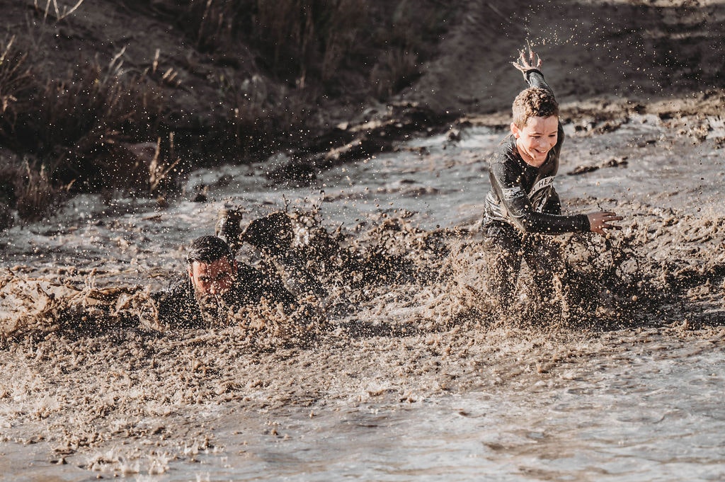 Two people covered in mud, sliding and splashing through a muddy area, having fun.