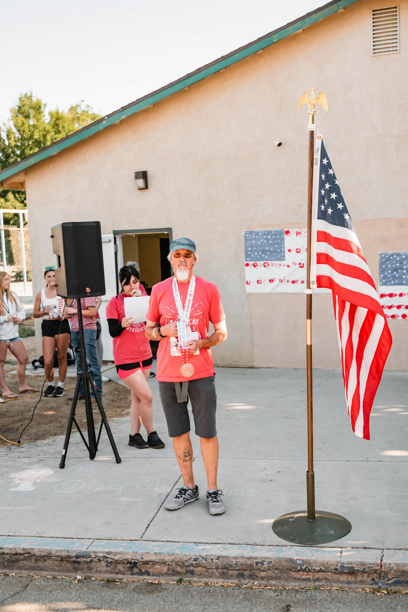 A man with medals stands next to an American flag, with a microphone and a group of people in the background.