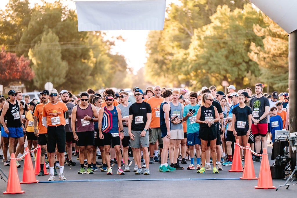 A group of people gathered at a starting line for a race, surrounded by orange cones, trees in the background.