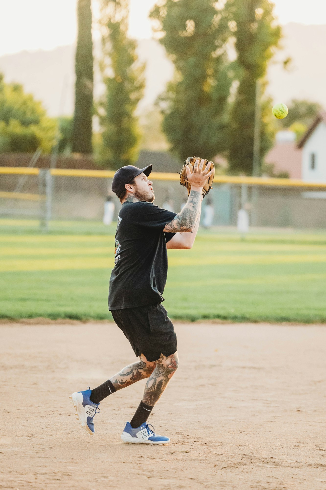 A person playing softball, catching a ball, wearing a cap, sports attire, and tattoos visible. Outdoor setting during sunset.