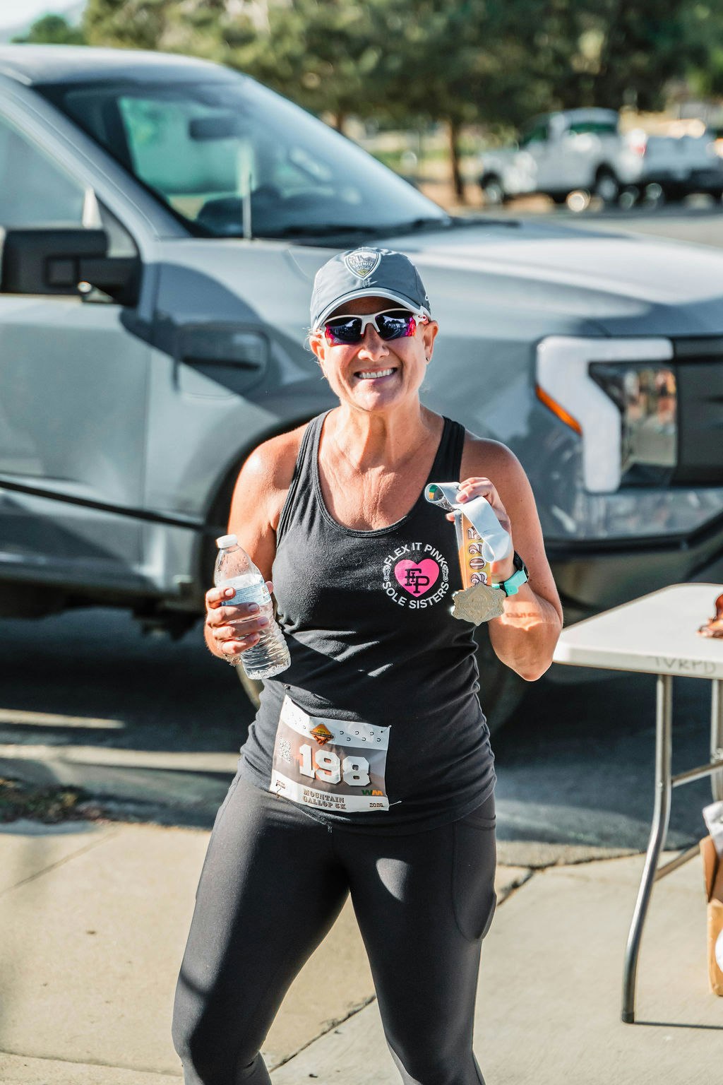 A smiling runner holding a water bottle and a medal, wearing sunglasses and a cap, stands next to a silver vehicle with trees in the background.