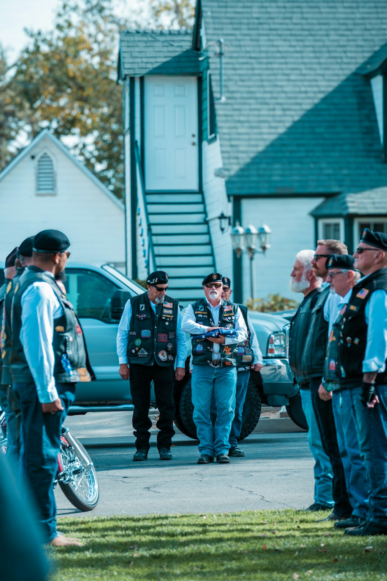 A group of people in vests stand outdoors in a semi-circle, one holding a folded flag, in front of houses.