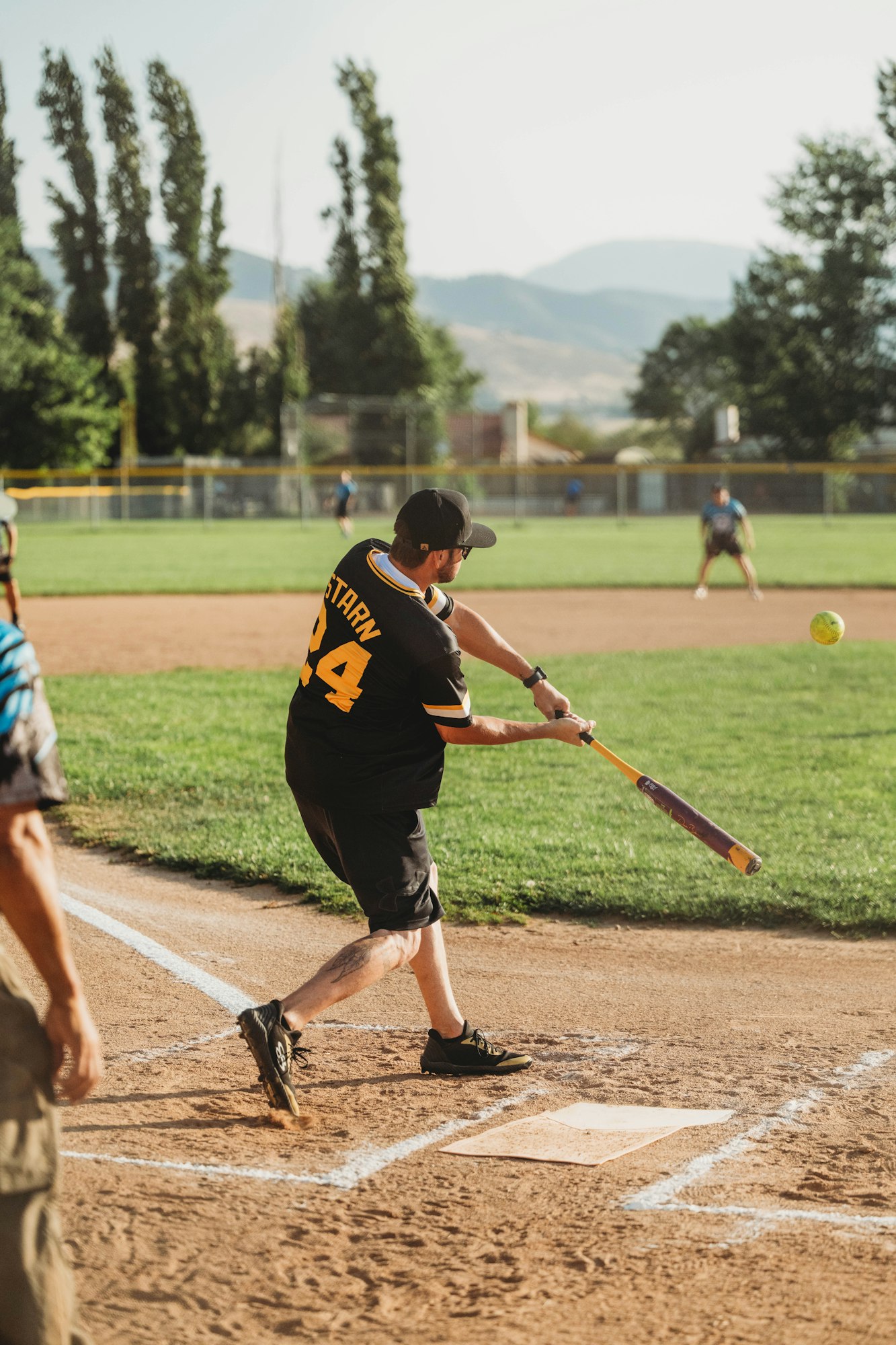 A batter swings at a ball during a softball game, with a fence and trees in the background.