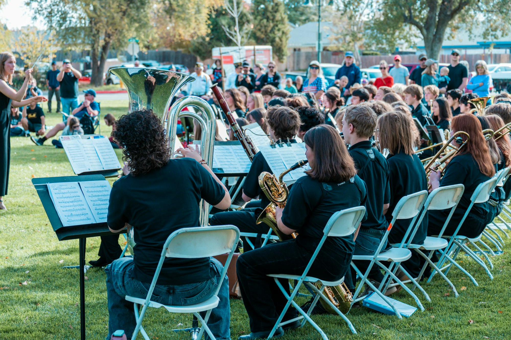 A band performs outdoors, with musicians playing instruments, sheet music displayed, and an audience in the background.