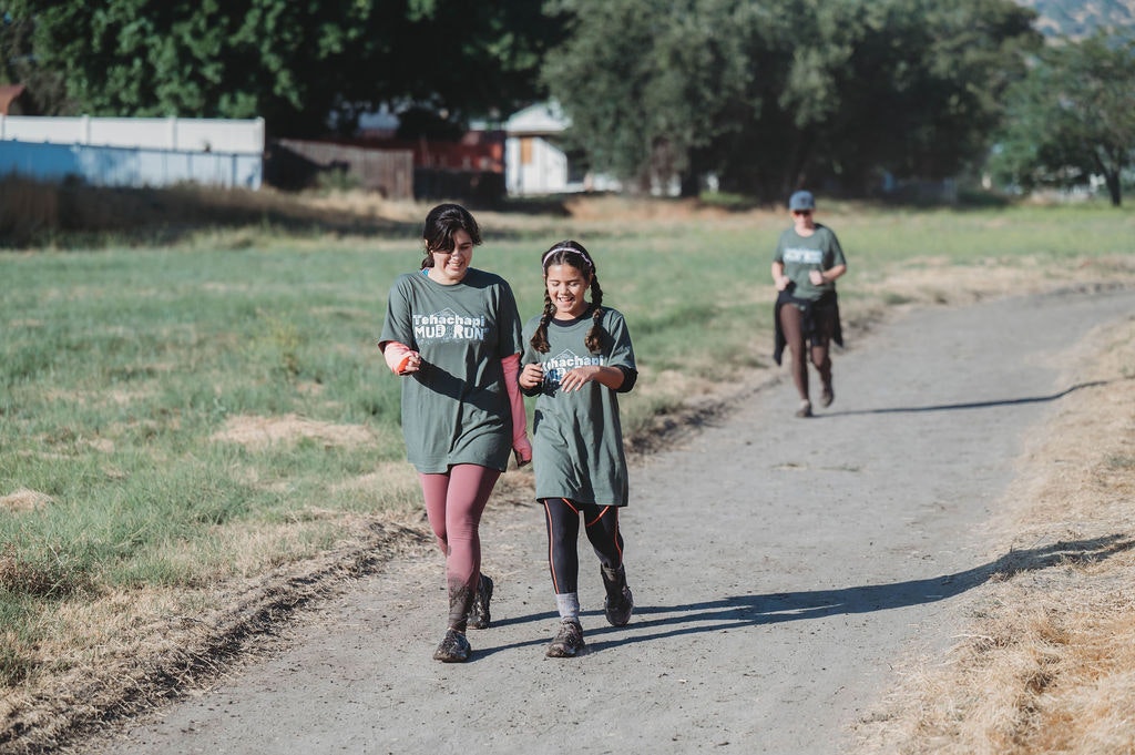 People walking in a mud run event, wearing green T-shirts and leggings on a grassy path.