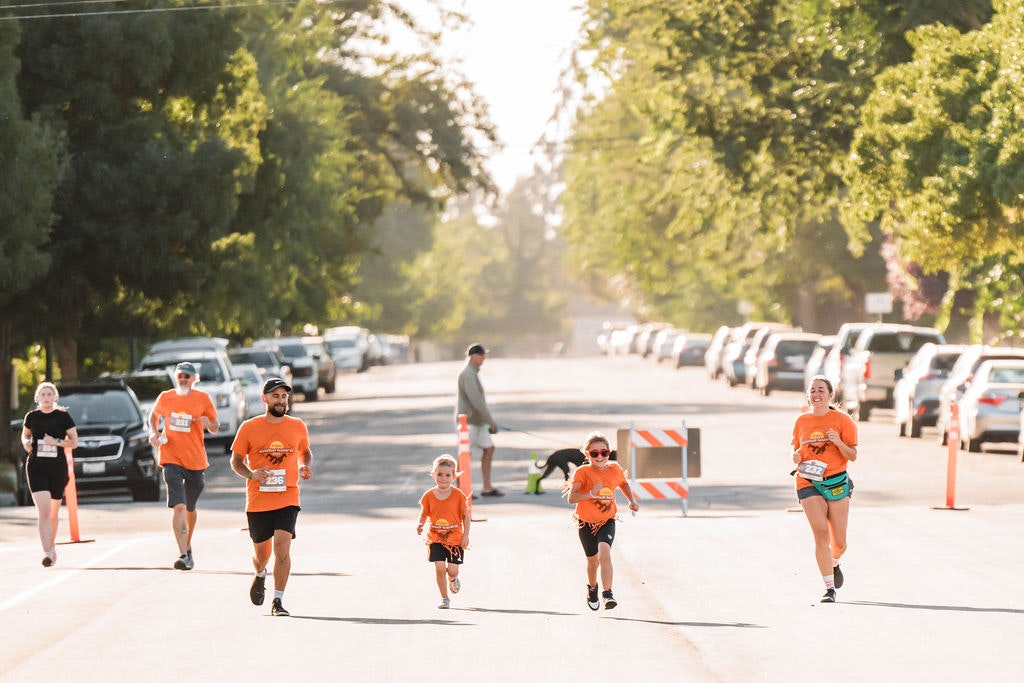 A group of people, including kids, running on a tree-lined street during a sunny day, wearing matching orange shirts and race numbers.