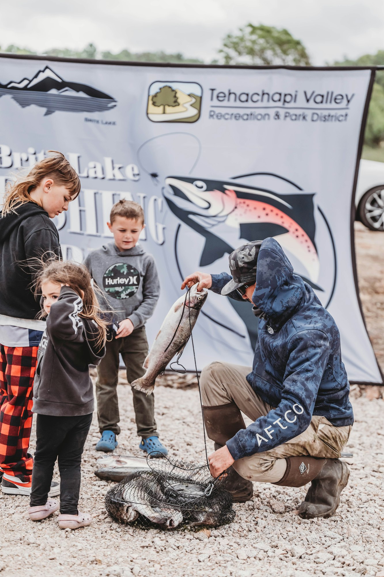 A group of people with fishing gear examine caught fish in front of a Tehachapi Valley fishing event banner.