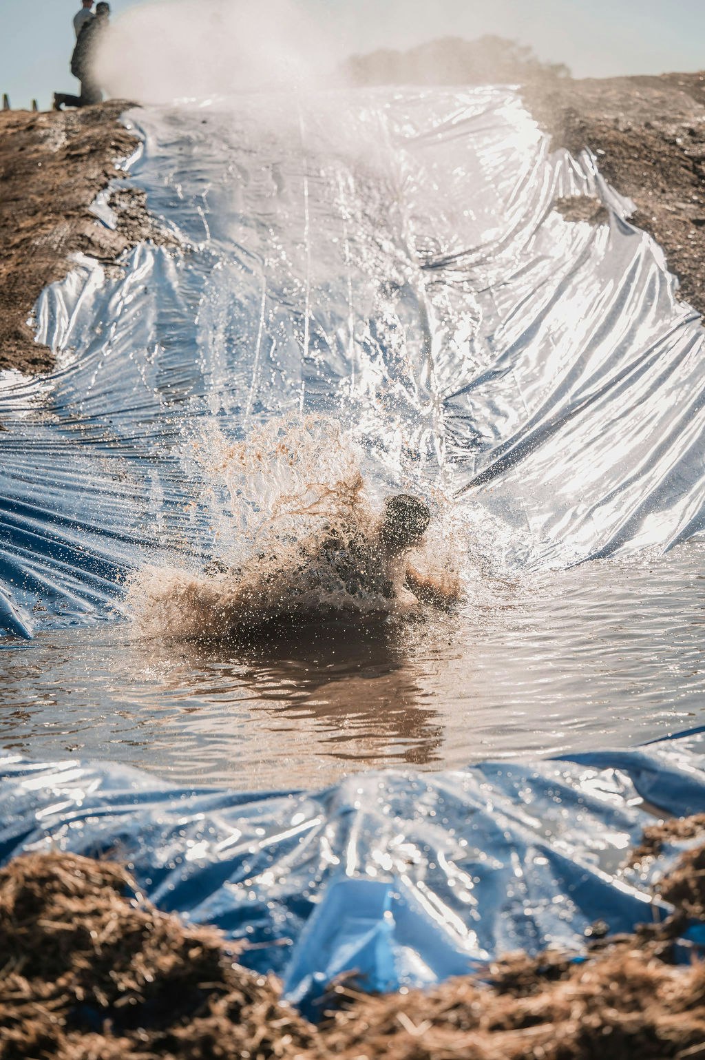 A person sliding down a muddy, wet plastic sheet, splashing into water at the bottom.