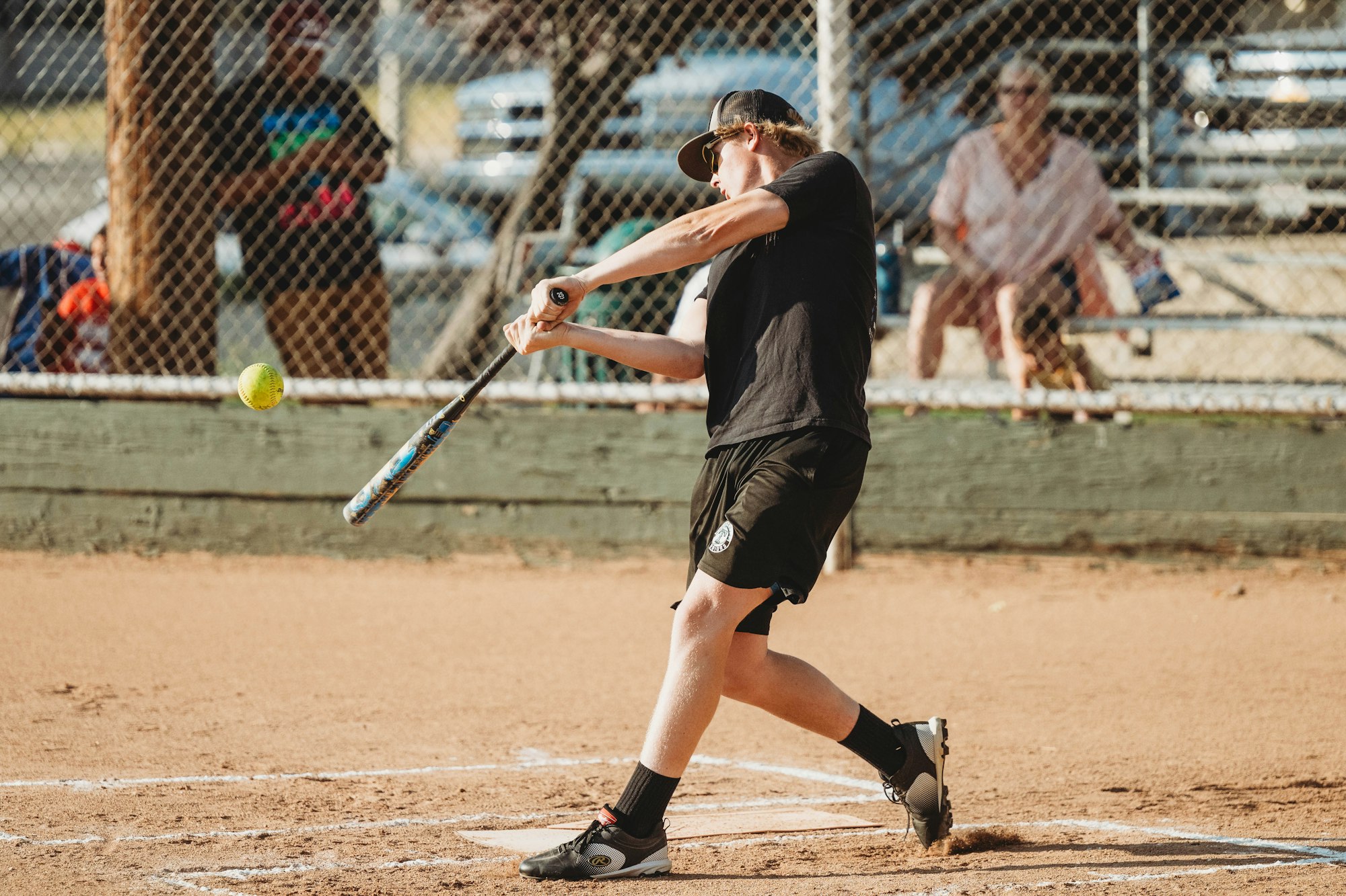 A person swinging a bat at a softball on a dusty field, with spectators in the background.