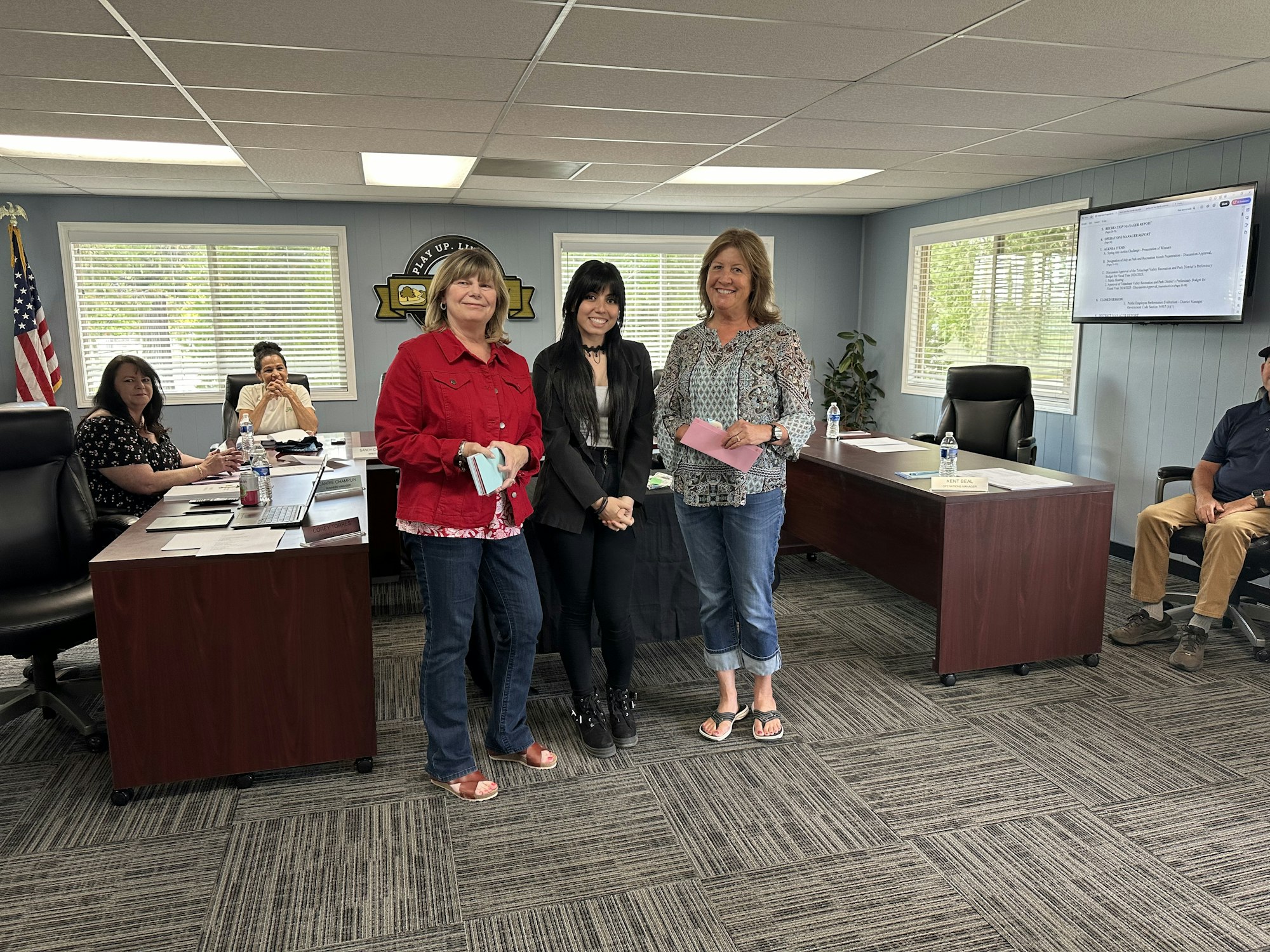 Three women standing with papers, smiling in an office setting, meeting in the background.