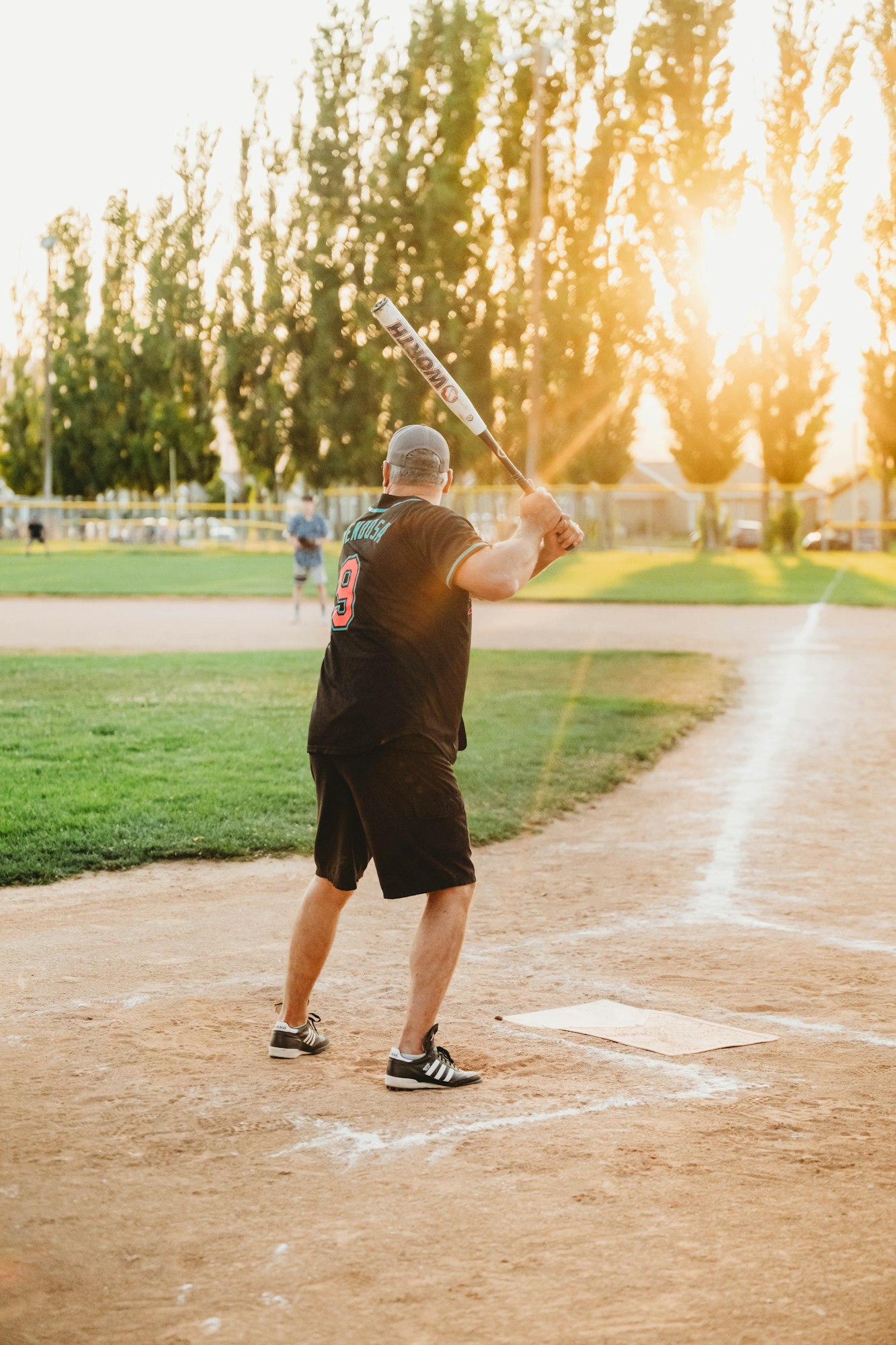 A person playing baseball, ready to bat, with trees and sunset in background.