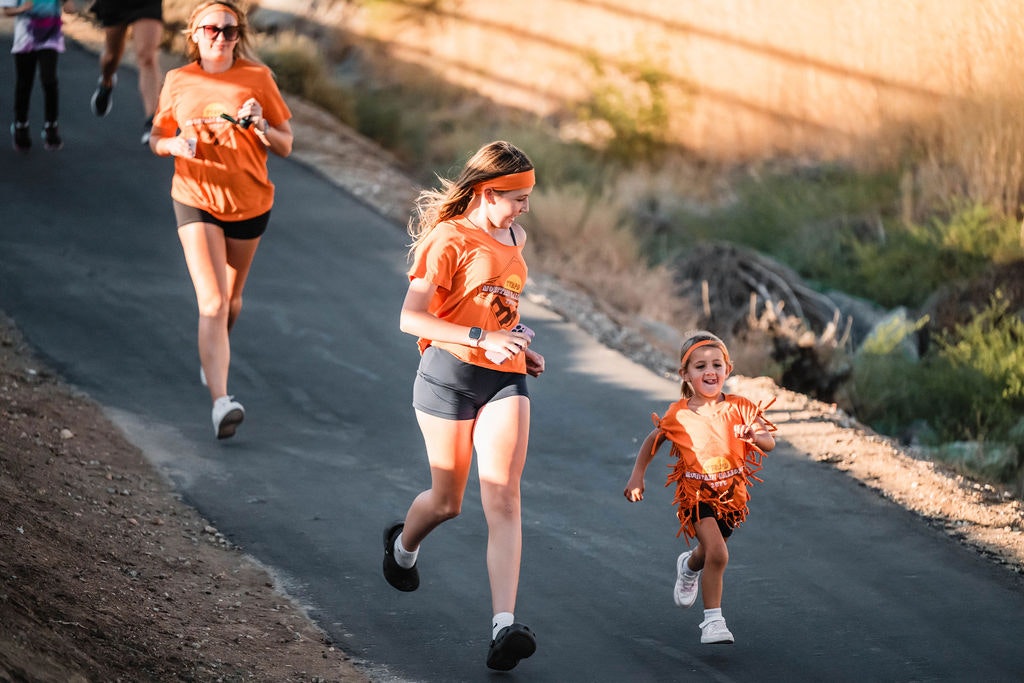 A group of people in orange shirts running on a paved path, with a child joyfully leading the way.