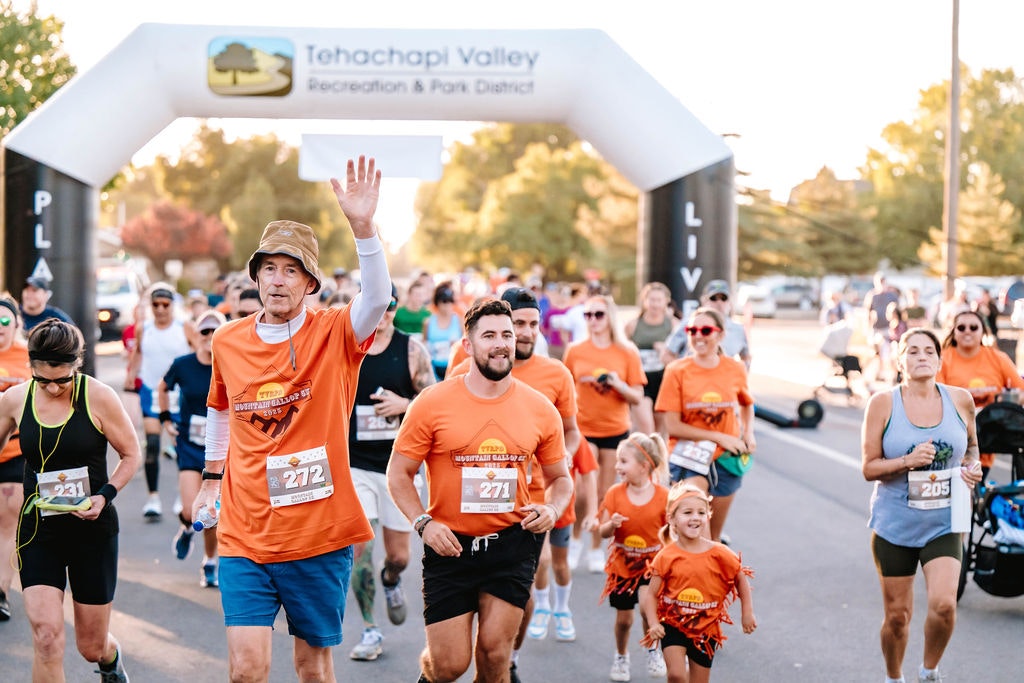 A group of people running in an outdoor race, wearing orange shirts with numbers, under an archway.