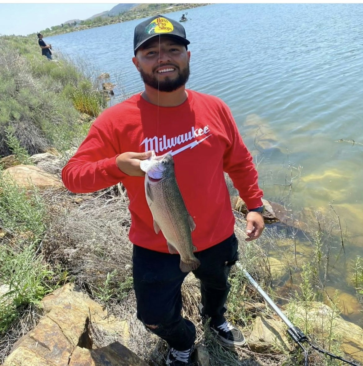 A person with a fish by a lake, a fishing rod, wearing a red shirt and cap. Another person is in the background, also fishing.