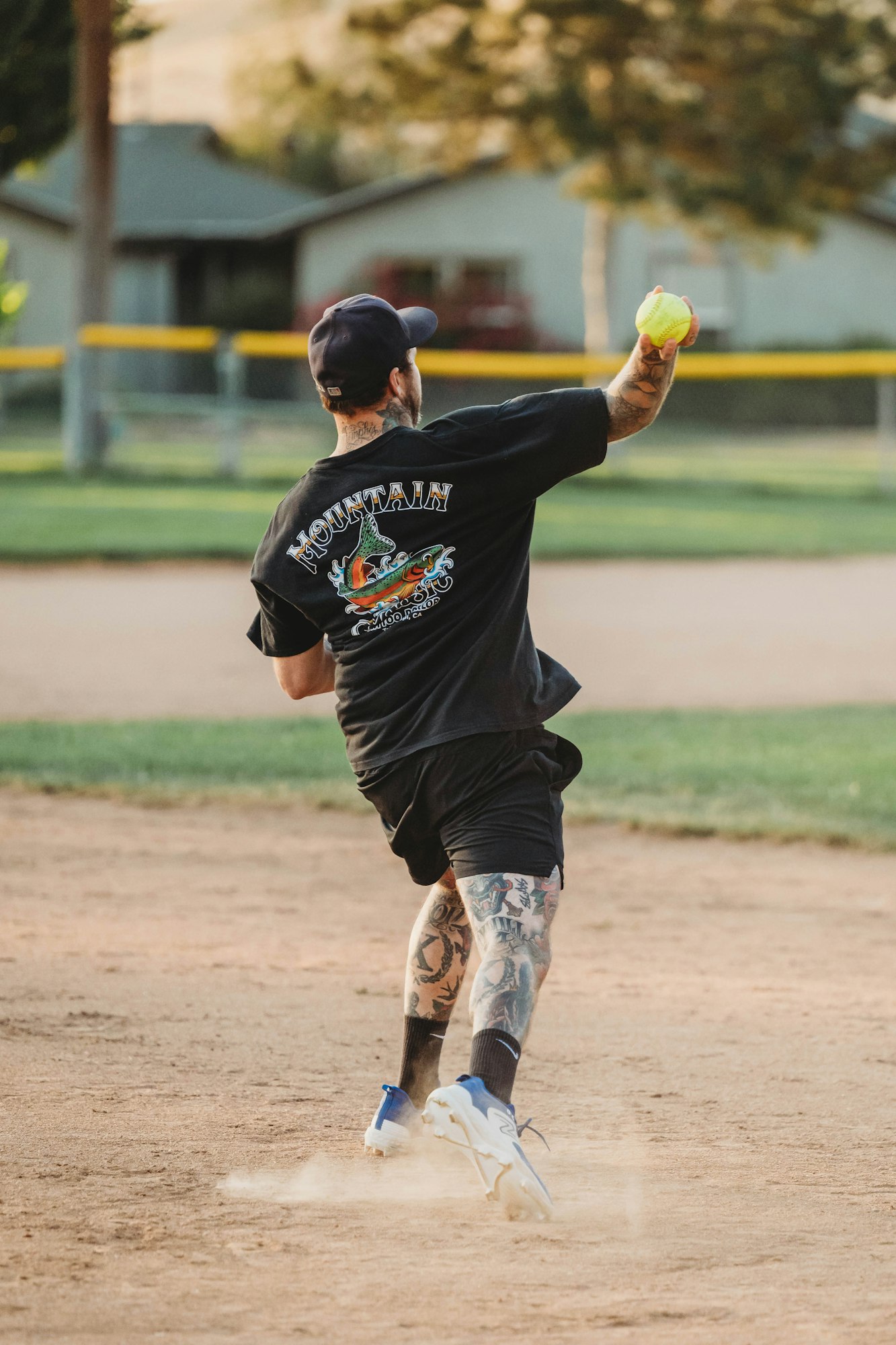 A person pitching a softball, tattooed arms, cap on, dust kicking up.