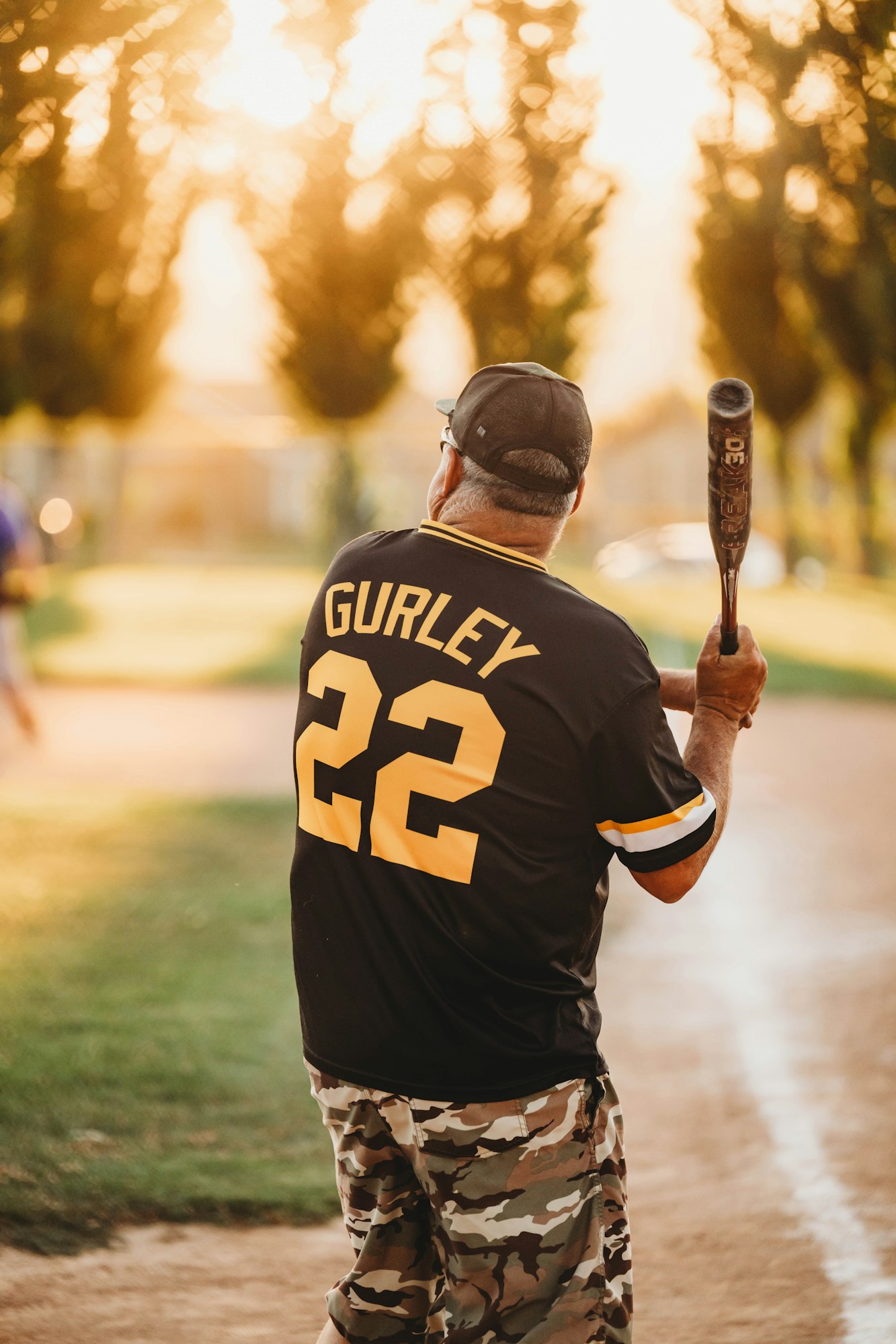 A person in a "GURLEY 22" jersey holds a baseball bat, with a sunset and park setting in the background.