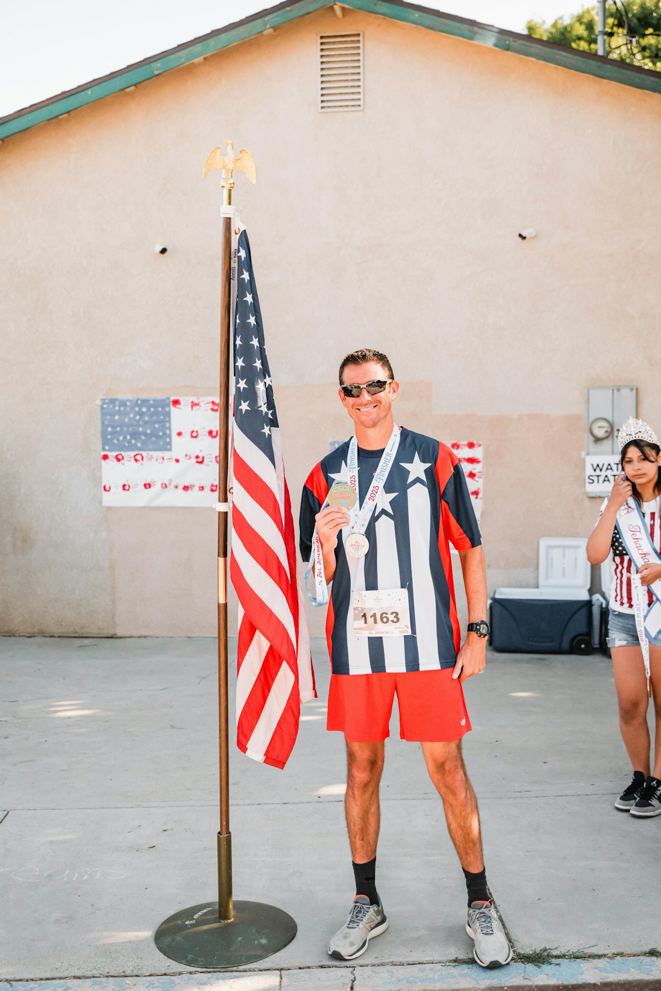 Man in star-striped outfit holding a medal, standing next to a U.S. flag. Another person in background with a sash and tiara.