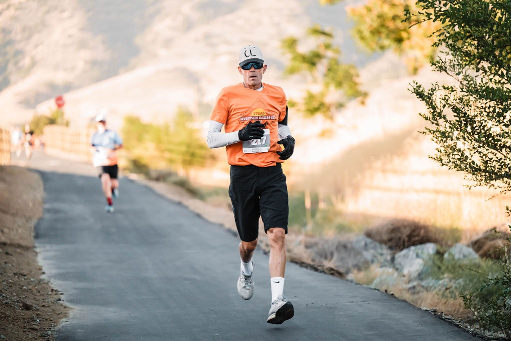 A person is running on a paved pathway outdoors, wearing an orange shirt and sunglasses, with another runner in the background.