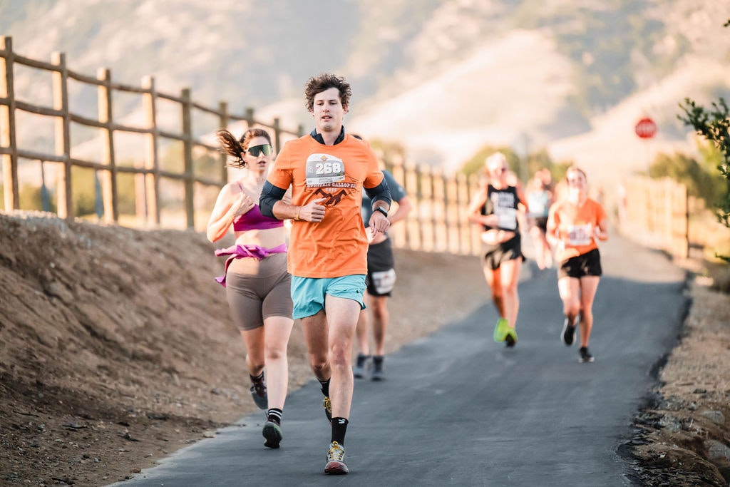 People running on a paved path during a race, with mountains and a wooden fence in the background.