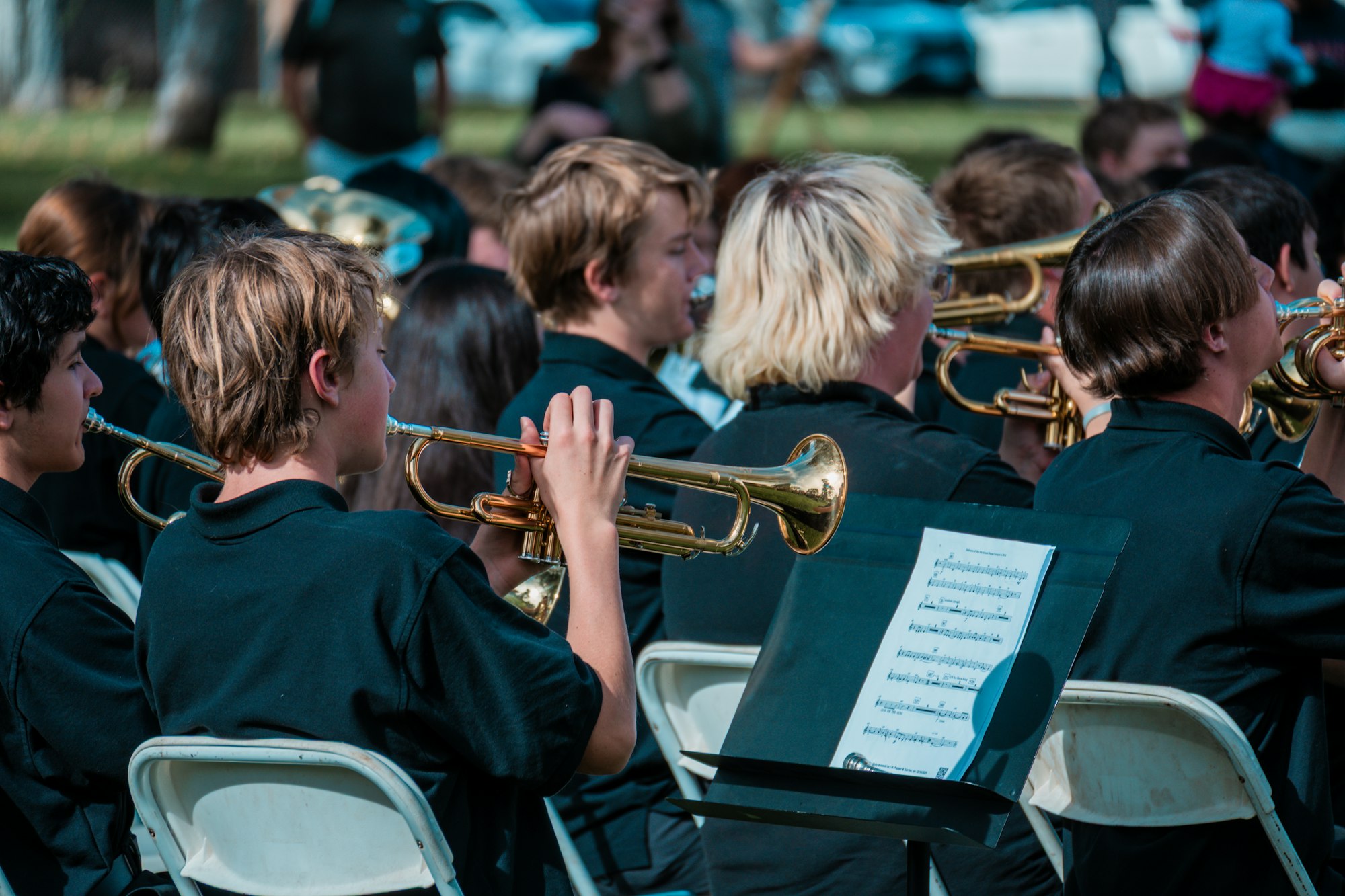 A group of musicians in black shirts playing trumpets outdoors with sheet music visible.