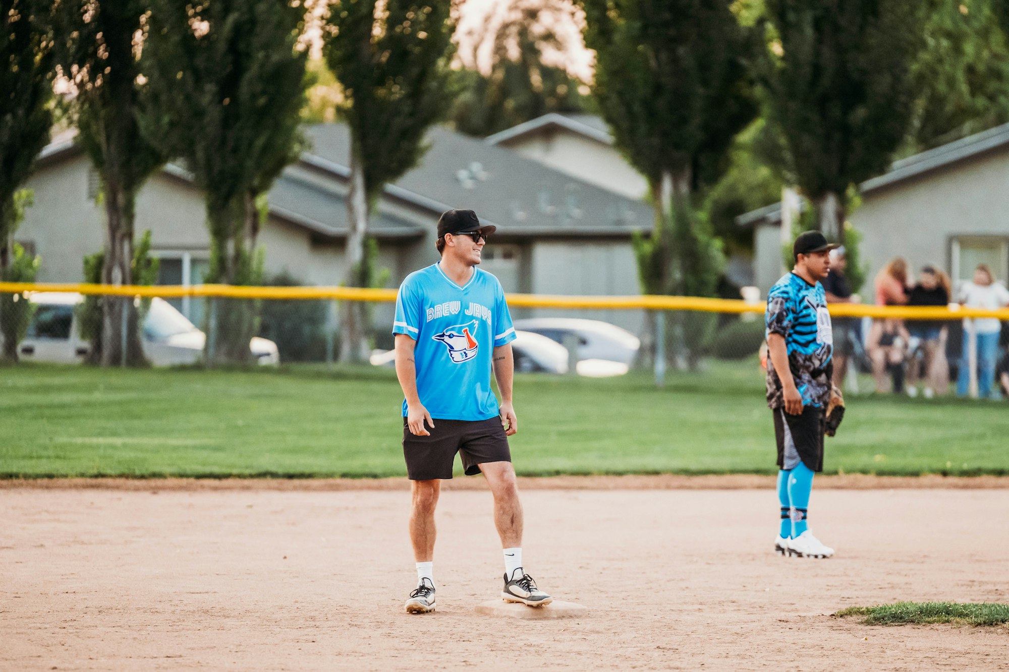 Two people playing softball on a field with trees in the background and a yellow fence.