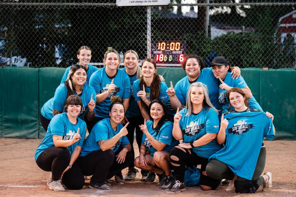 A women's softball team wearing blue shirts posing on a field, celebrating a win with smiles and hand gestures.