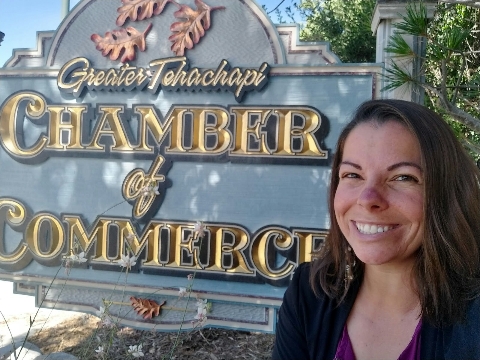 The image shows a smiling woman standing by the Greater Tehachapi Chamber of Commerce sign.