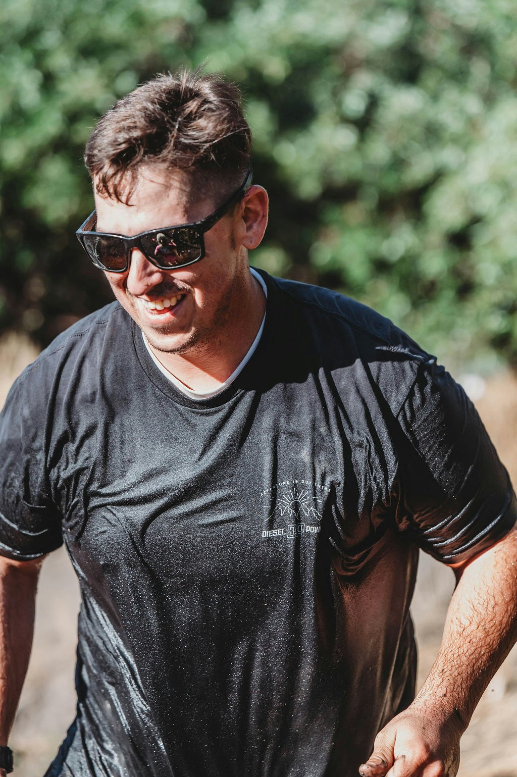 A man wearing sunglasses and a wet T-shirt smiles outdoors.