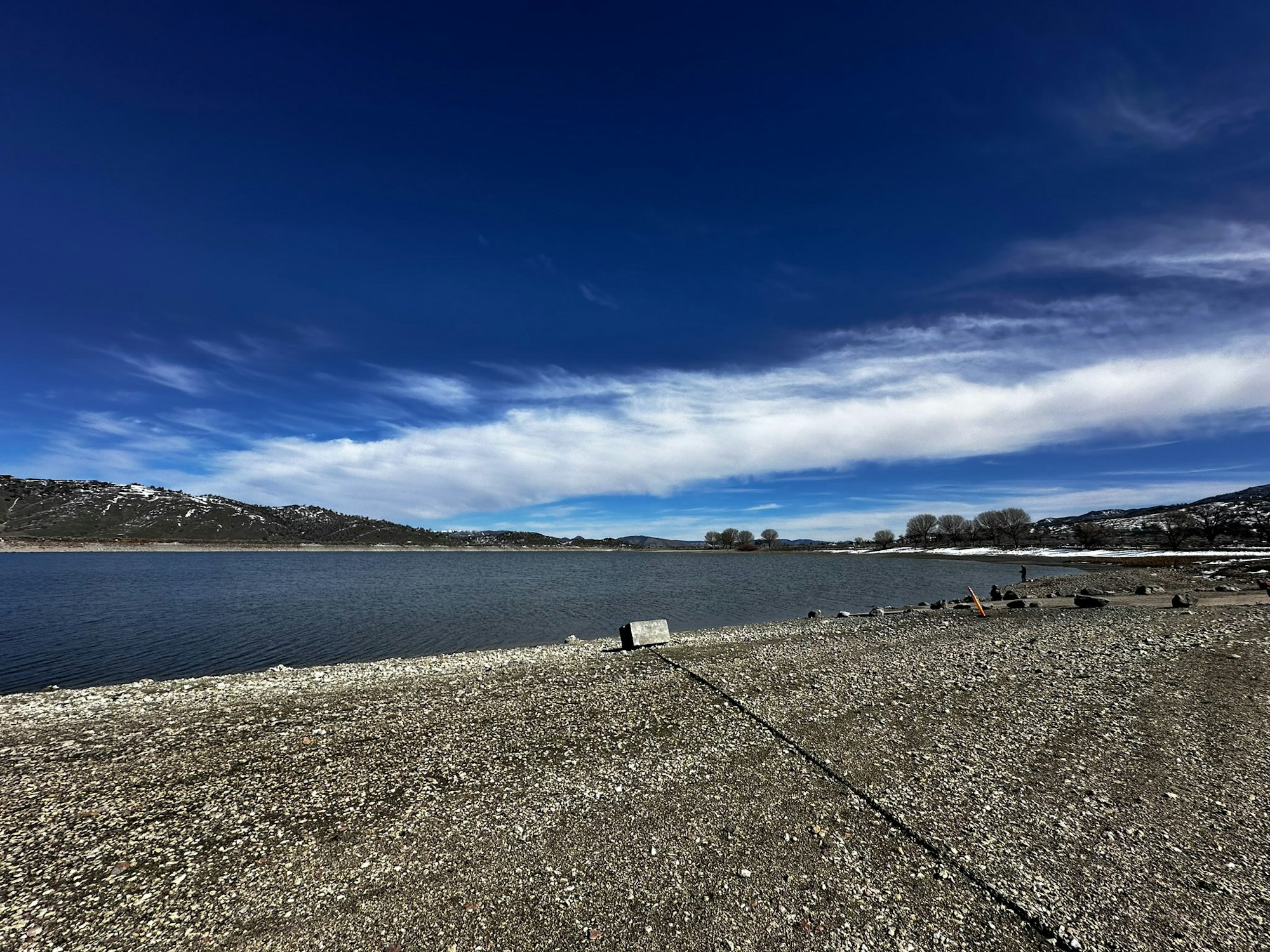 A serene lake with a pebbled shore, against a backdrop of hills under a vast, blue sky with wispy clouds.