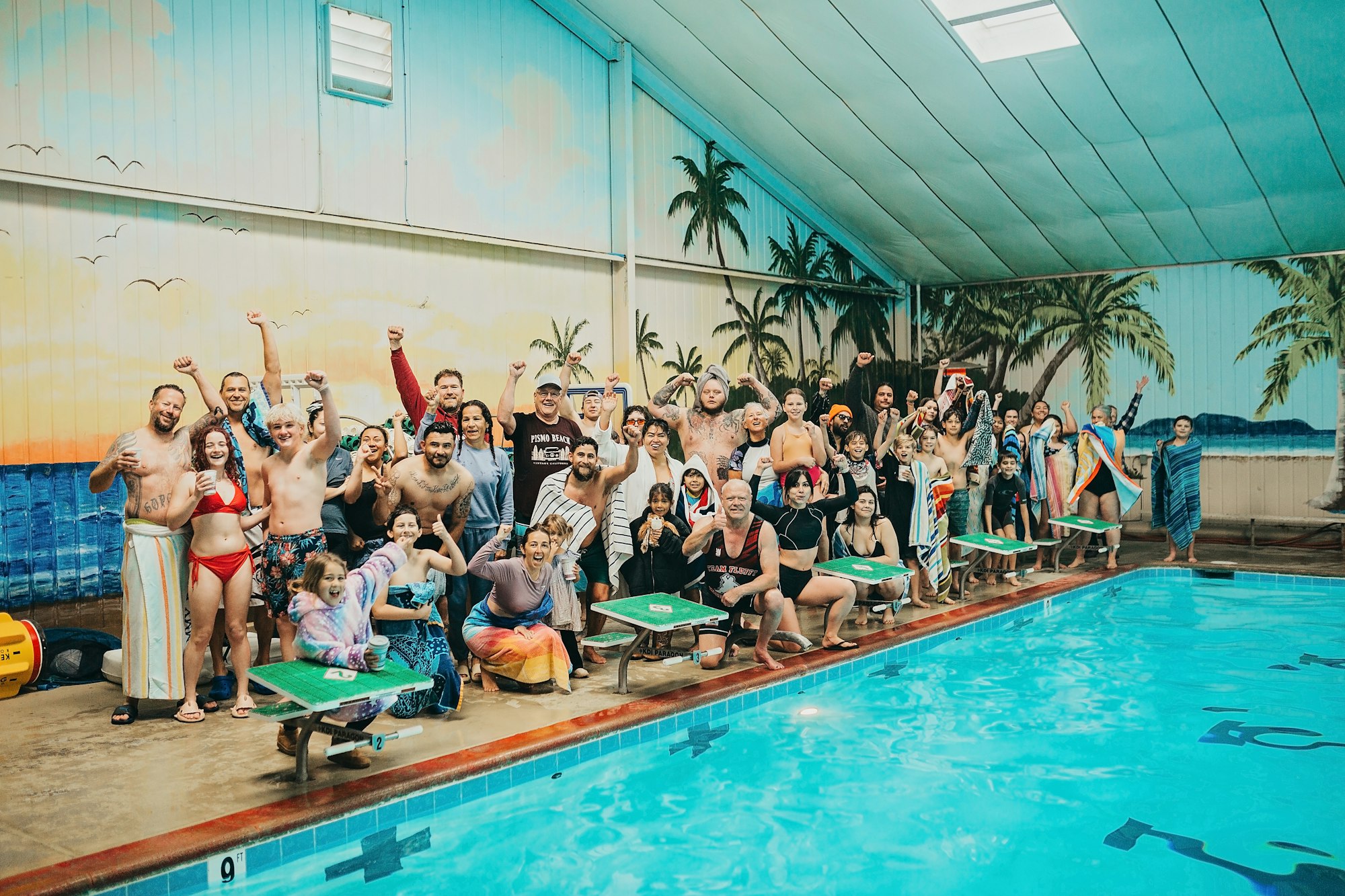 A large group of people posing happily by an indoor pool with a tropical mural background.